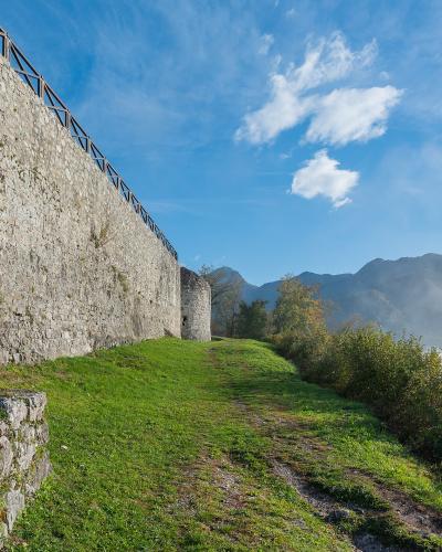 A stone fortress wall with a modern wooden railing runs along a grassy slope, overlooking a valley with mountains and low-lying clouds in the background.