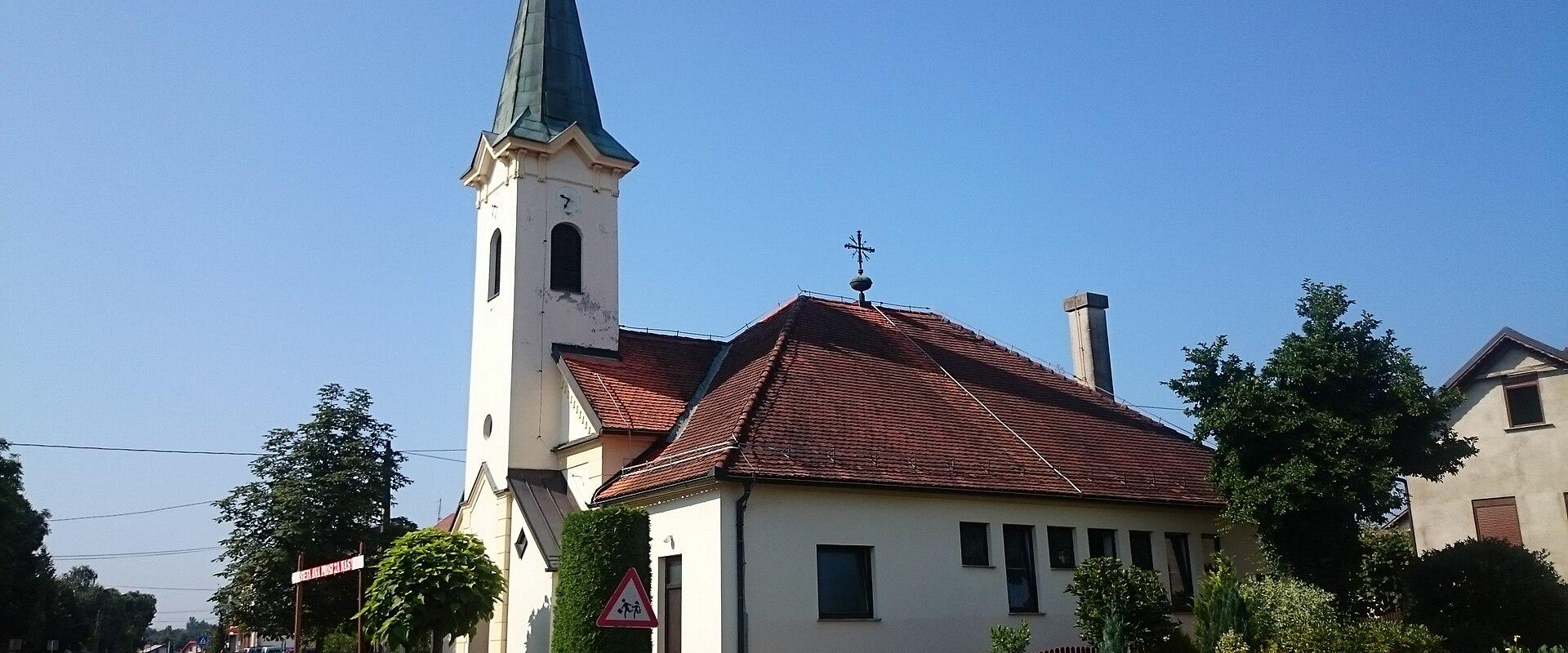 A white church with a tall, slender steeple topped by a green spire and a cross, situated next to a red-tiled building and a wooden fence along a paved road under a clear blue sky.