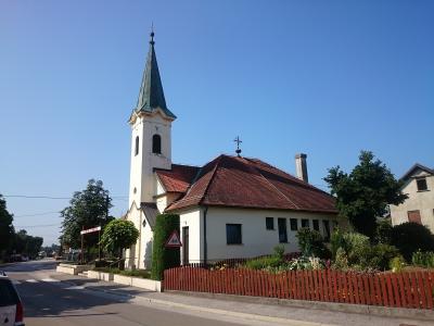 A white church with a tall, slender steeple topped by a green spire and a cross, situated next to a red-tiled building and a wooden fence along a paved road under a clear blue sky.