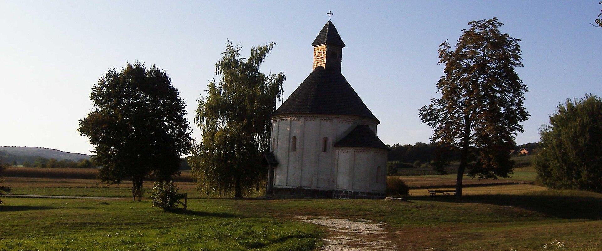 A white church with a dark conical roof stands in a grassy field, surrounded by trees and a gravel path, under a clear blue sky.