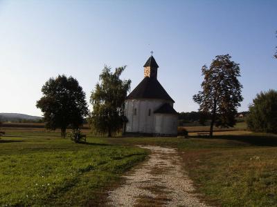 A white church with a dark conical roof stands in a grassy field, surrounded by trees and a gravel path, under a clear blue sky.