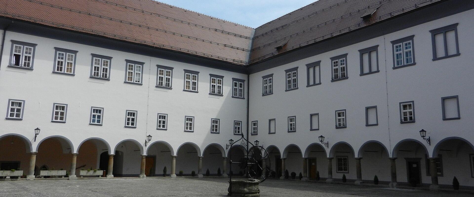 The image shows a large, paved courtyard surrounded by two-story white buildings with arched arcades on the ground floor and rows of rectangular windows above, featuring a central stone well or fountain structure.