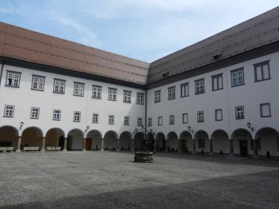The image shows a large, paved courtyard surrounded by two-story white buildings with arched arcades on the ground floor and rows of rectangular windows above, featuring a central stone well or fountain structure.