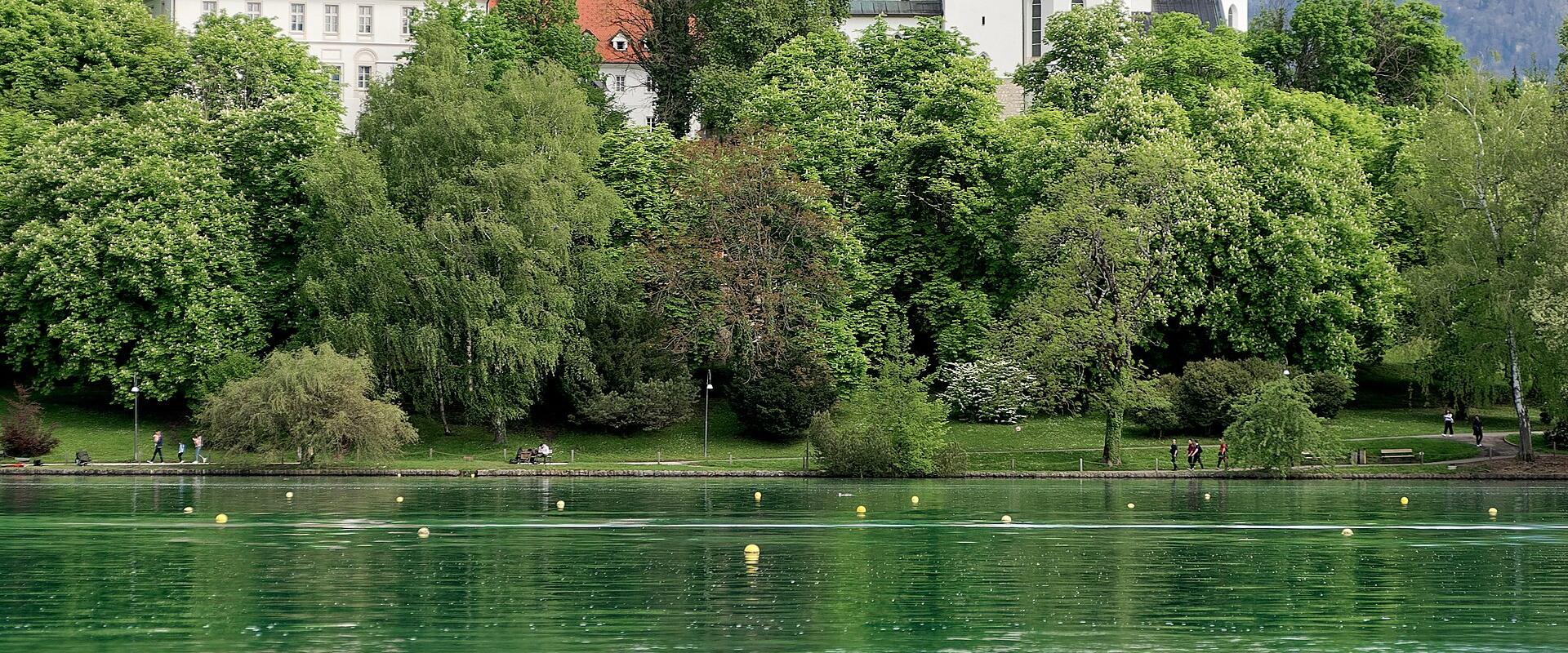 A white church with a tall, slender spire and distinctive patterned roof tiles stands on the shore of a green lake, with a rocky cliff face to the left and mountains in the background.