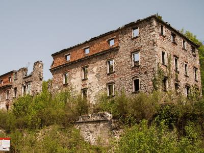 The image shows the stone and brick ruins of a large, multi-story medieval castle structure situated on a grassy, overgrown hillside under a clear sky.