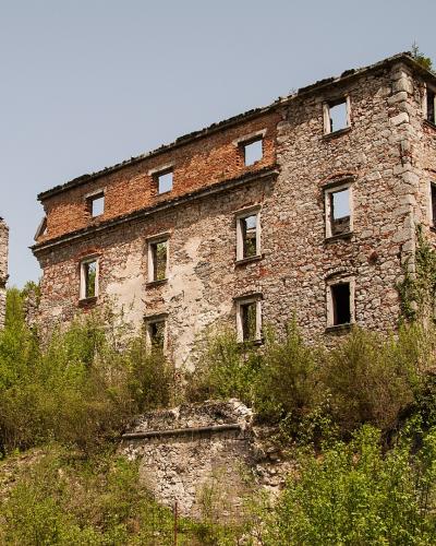 The image shows the stone and brick ruins of a large, multi-story medieval castle structure situated on a grassy, overgrown hillside under a clear sky.