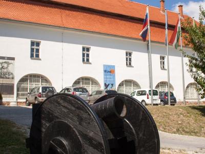 A white building with a red tiled roof, arched windows, and flags (including Slovenian and Hungarian) flying outside, with a large black wooden cannon in the foreground.