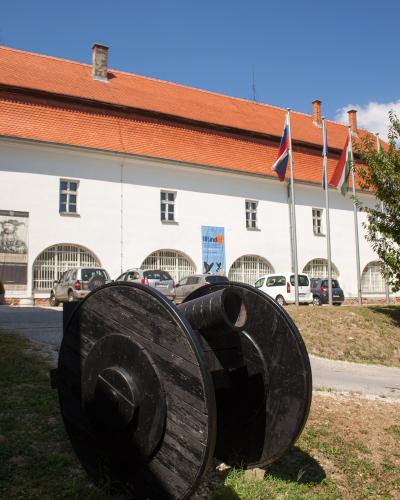 A white building with a red tiled roof, arched windows, and flags (including Slovenian and Hungarian) flying outside, with a large black wooden cannon in the foreground.
