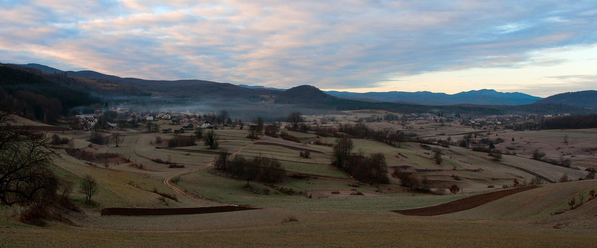 A panoramic landscape view showing rolling green hills, scattered rural houses, and a misty valley, with a prominent hill in the center distance, captured during twilight or early morning.