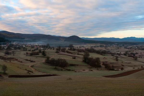 A panoramic landscape view showing rolling green hills, scattered rural houses, and a misty valley, with a prominent hill in the center distance, captured during twilight or early morning.