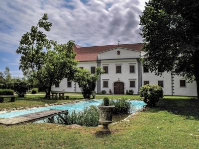 A white building with a red-tiled roof stands behind a small pond, surrounded by green trees and grass under a cloudy sky.