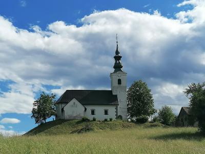 A white church with a tall, dark-spired bell tower stands atop a grassy hill under a blue sky with white clouds.