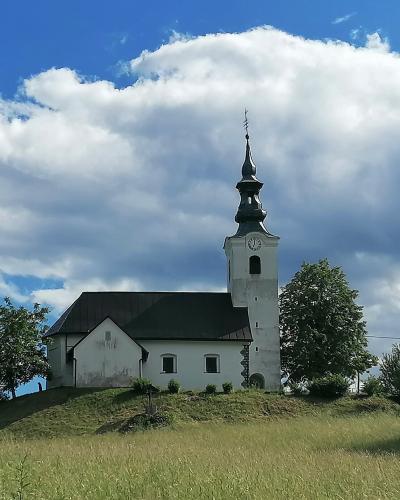 A white church with a tall, dark-spired bell tower stands atop a grassy hill under a blue sky with white clouds.