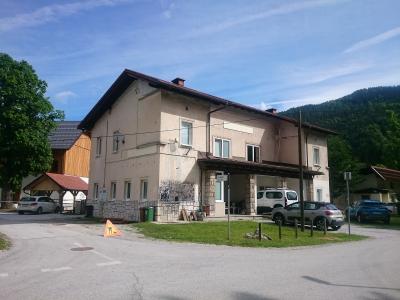 A two-story beige building with a covered entrance supported by stone pillars, situated at a road junction with parked cars and a forested hill in the background.