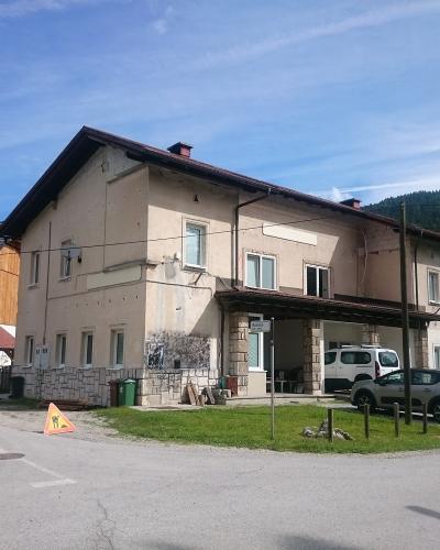 A two-story beige building with a covered entrance supported by stone pillars, situated at a road junction with parked cars and a forested hill in the background.