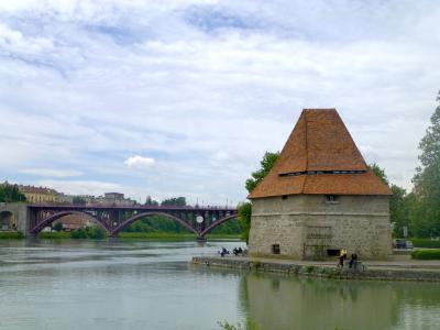 A stone pentagonal water tower with a tiled pyramidal roof stands on the bank of a river, with a red arched bridge spanning the water in the background under a cloudy sky.