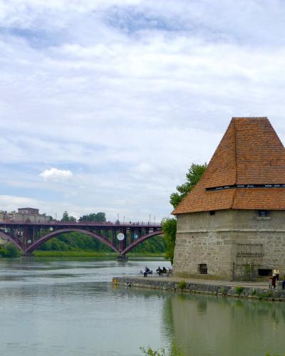 A stone pentagonal water tower with a tiled pyramidal roof stands on the bank of a river, with a red arched bridge spanning the water in the background under a cloudy sky.