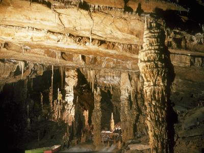 A caver wearing a red helmet and holding a camera is squeezing through a narrow, rocky cave passage.