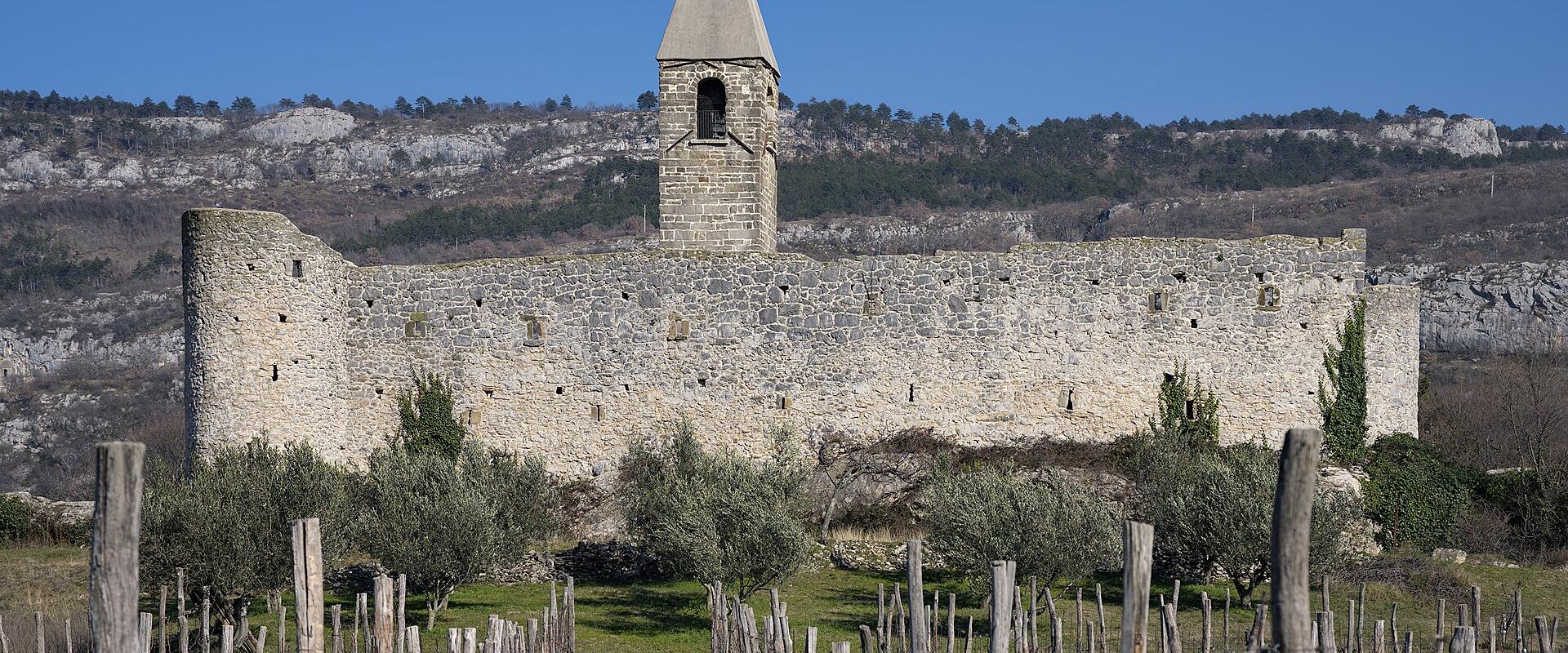 A fortified stone church with a prominent bell tower and crenellated walls, situated on a hillside with vineyards in the foreground and limestone cliffs in the background.