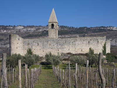 A fortified stone church with a prominent bell tower and crenellated walls, situated on a hillside with vineyards in the foreground and limestone cliffs in the background.
