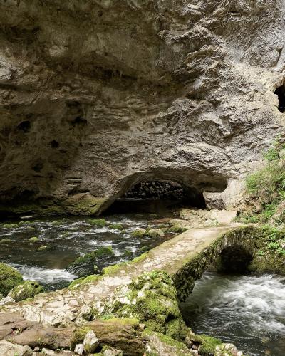 A low, moss-covered stone footbridge spanning a rushing stream within a rocky gorge, with a large natural cave opening visible in the background cliff face.