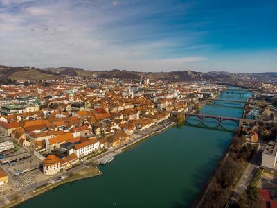 An aerial view of a European city with orange-roofed buildings along a wide river, featuring a prominent round tower in the foreground and multiple bridges crossing the water.