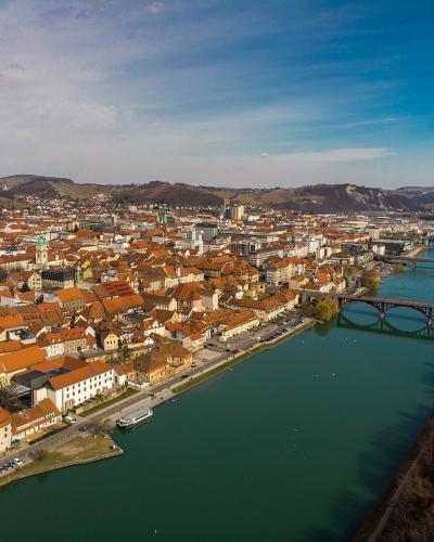 An aerial view of a European city with orange-roofed buildings along a wide river, featuring a prominent round tower in the foreground and multiple bridges crossing the water.