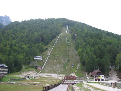 A large ski flying hill with a grassy in-run and landing slope, surrounded by forested hills, with buildings at the base and a mountain peak visible in the background.
