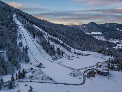 An aerial view of a large ski jumping complex in a snowy valley, featuring a prominent large hill on the left, smaller hills, and a distinctive circular modern building in the foreground.