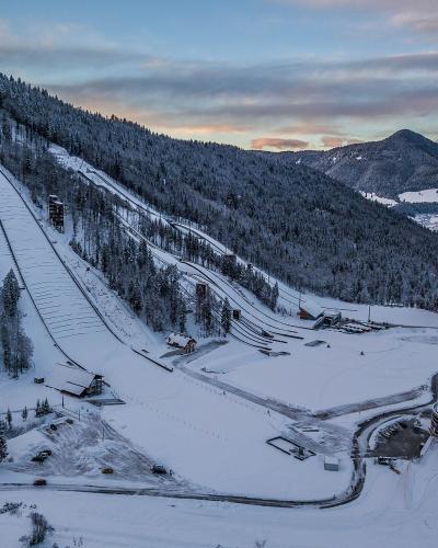 An aerial view of a large ski jumping complex in a snowy valley, featuring a prominent large hill on the left, smaller hills, and a distinctive circular modern building in the foreground.