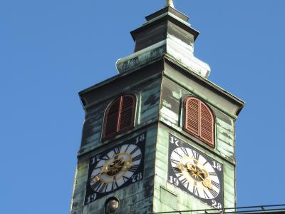 A close-up view of the clock tower of a historic building, featuring a green copper roof, two visible clock faces with Roman numerals and golden hands, arched louvered windows, and a golden trident finial atop the spire, set against a clear blue s