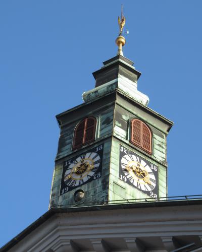 A close-up view of the clock tower of a historic building, featuring a green copper roof, two visible clock faces with Roman numerals and golden hands, arched louvered windows, and a golden trident finial atop the spire, set against a clear blue s
