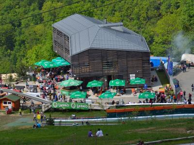 A large, modern wooden building with a grey roof sits on a hillside, surrounded by green trees and a crowded outdoor terrace with green umbrellas bearing the "LASKO" logo, with a toboggan run visible in the foreground.