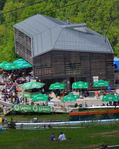 A large, modern wooden building with a grey roof sits on a hillside, surrounded by green trees and a crowded outdoor terrace with green umbrellas bearing the "LASKO" logo, with a toboggan run visible in the foreground.