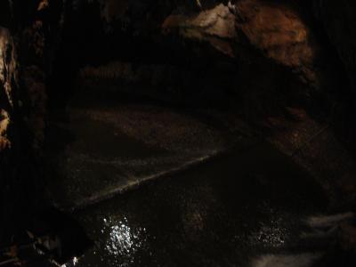 A dark underground cave chamber featuring a flowing river with a small weir or dam, illuminated by artificial lights, with a walkway visible along the upper left wall.