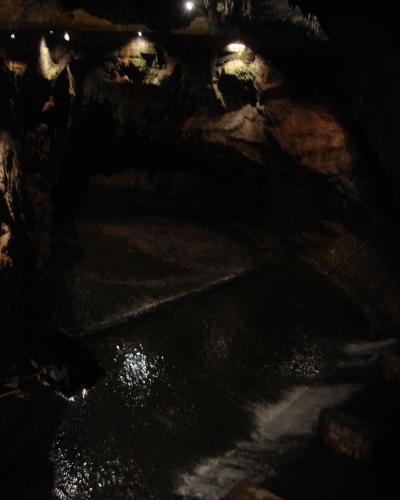 A dark underground cave chamber featuring a flowing river with a small weir or dam, illuminated by artificial lights, with a walkway visible along the upper left wall.