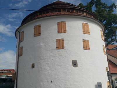 A low-angle shot of a white, cylindrical medieval tower with wooden shutters on its windows and a distinctive conical tiled roof, situated on a paved street with cars and a bench in the foreground under a blue sky.