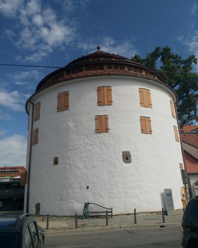 A low-angle shot of a white, cylindrical medieval tower with wooden shutters on its windows and a distinctive conical tiled roof, situated on a paved street with cars and a bench in the foreground under a blue sky.