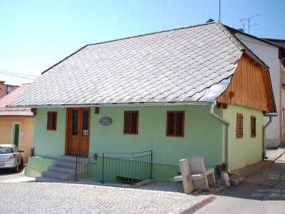 A single-story, light green house with a grey tiled roof and wooden shutters, situated on a cobblestone street corner with a silver car parked nearby and a stone bench in the foreground.