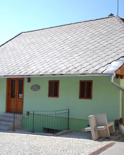 A single-story, light green house with a grey tiled roof and wooden shutters, situated on a cobblestone street corner with a silver car parked nearby and a stone bench in the foreground.