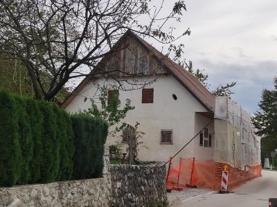 A white, two-story house with a steep gabled roof and wooden shutters, partially covered in scaffolding and protective sheeting for renovation, situated behind a stone wall and orange safety fencing.