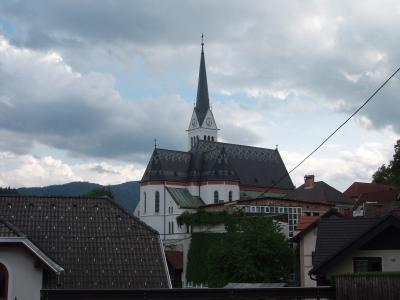 A white church with a tall, dark spire and a distinctive patterned roof stands in a town setting with other buildings and hills in the background.