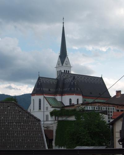 A white church with a tall, dark spire and a distinctive patterned roof stands in a town setting with other buildings and hills in the background.