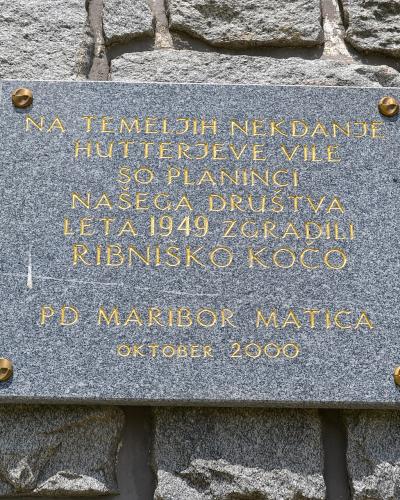 A close-up photograph of a grey granite commemorative plaque mounted on a rough stone wall, featuring gold lettering that reads "NA TEMELJIH NEKDANJE HUTTERJEVE VILE SO PLANINCI NAŠEGA DRUŠTVA LETA 1949 ZGRADILI RIBNIŠKO KOČO" and "PD MARIBOR MATICA 