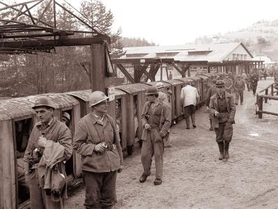 A black-and-white photograph showing a group of miners in work clothes and helmets standing next to a line of wooden mine carts on a track, with a large industrial headframe structure overhead and a building in the background.