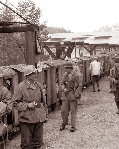 A black-and-white photograph showing a group of miners in work clothes and helmets standing next to a line of wooden mine carts on a track, with a large industrial headframe structure overhead and a building in the background.