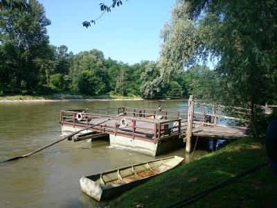 A cable ferry docked at a wooden pier on a wide, green river, with a small boat beached on the grassy bank in the foreground.