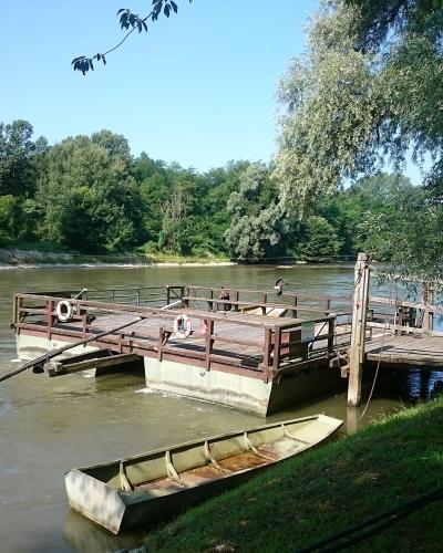 A cable ferry docked at a wooden pier on a wide, green river, with a small boat beached on the grassy bank in the foreground.