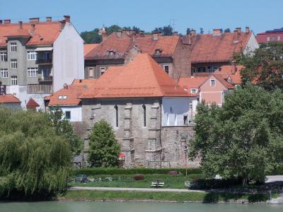A stone building with a red-tiled roof, situated along a riverbank with greenery and other buildings in the background; no visible signage or identifying text.