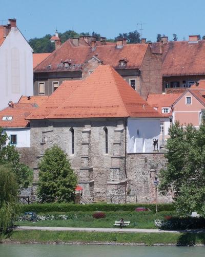 A stone building with a red-tiled roof, situated along a riverbank with greenery and other buildings in the background; no visible signage or identifying text.